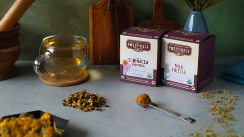 Two botanical tea boxes among piles of loose-leaf botanicals, serving trays, a mortar and pestle, and a mug of tea.