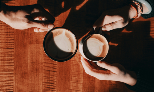 Two cups of coffee being held by hands over a wood table. 