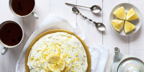 2 cups of coffee, a lemon pie topped with white cream, lemon slices, and lemon peel, and a white plate with lemon wedges sit on a white table. There is also a teapot and two spoons. 