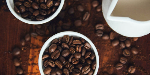 Two white bowls full of roasted coffee beans on a wooden table surrounded by loose coffee beans and a white pitcher of milk