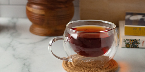 A glass cup of herbal tea sits on a woven coaster in front of some books and a mortar and pestle. 