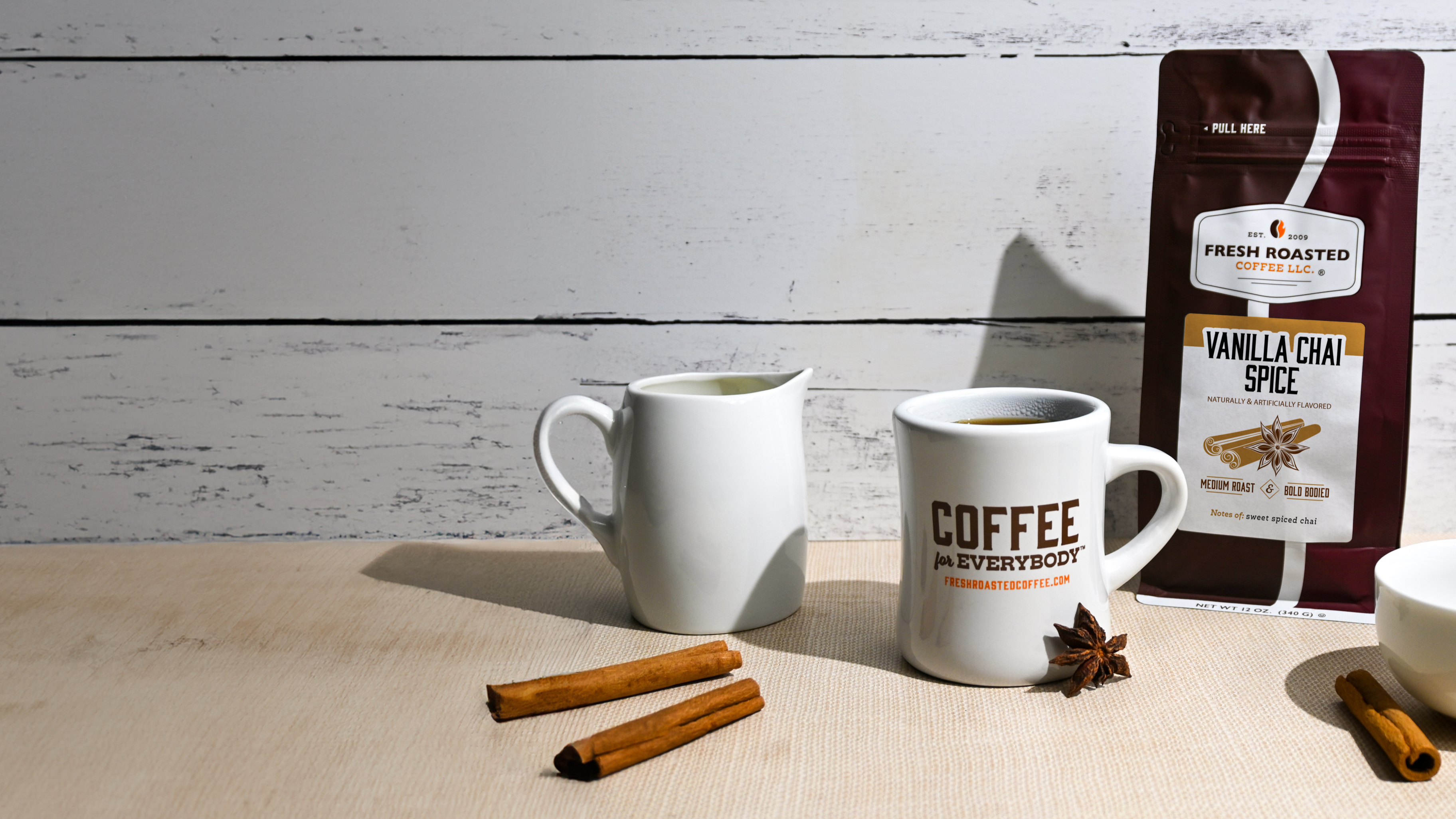 Vanilla Chai Spice Coffee in a Fresh Roasted Coffee brown and maroon bag with a white mug with "coffee for everybody" printed on the side. Also in image - a white milk pitcher, cinnamon sticks, and star anise against a wooden background, painted white. 